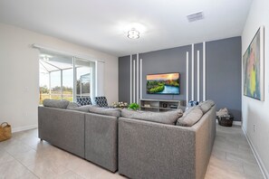 Ein modernes Wohnzimmer mit einer Eckcouch, einem flachbildfernseher an der Wand und einem großen Fenster mit Blick ins Freie.
