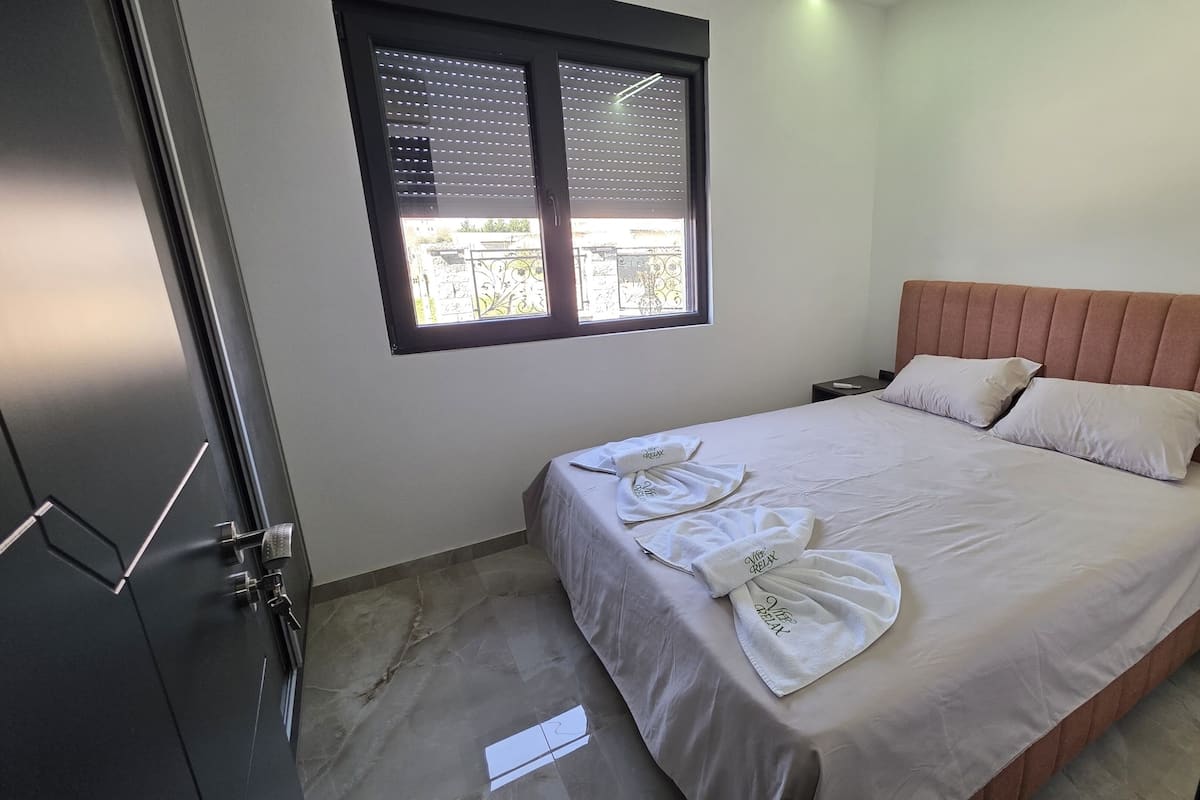 Une chambre à coucher avec un lit, des oreillers, une fenêtre avec des stores, une table de chevet et une porte.