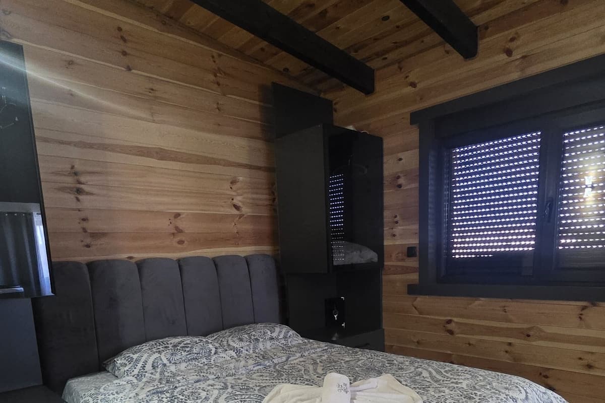 Une chambre avec un plafond en bois, un lit recouvert d’une couvre-lit à motifs, une tête de lit sombre, une table de chevet, une armoire noire et une fenêtre avec des stores.
