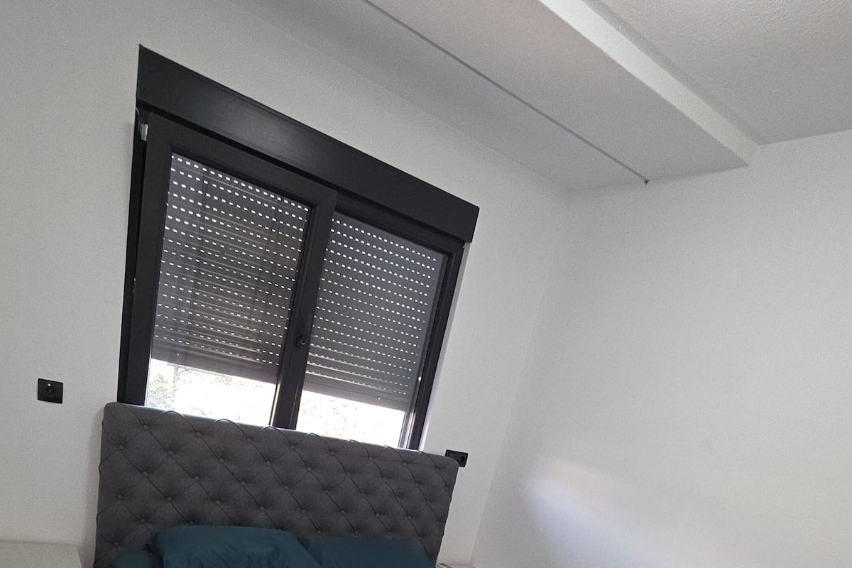 Une chambre à coucher avec un lit, une fenêtre avec des stores, une table de chevet et une prise électrique au mur.