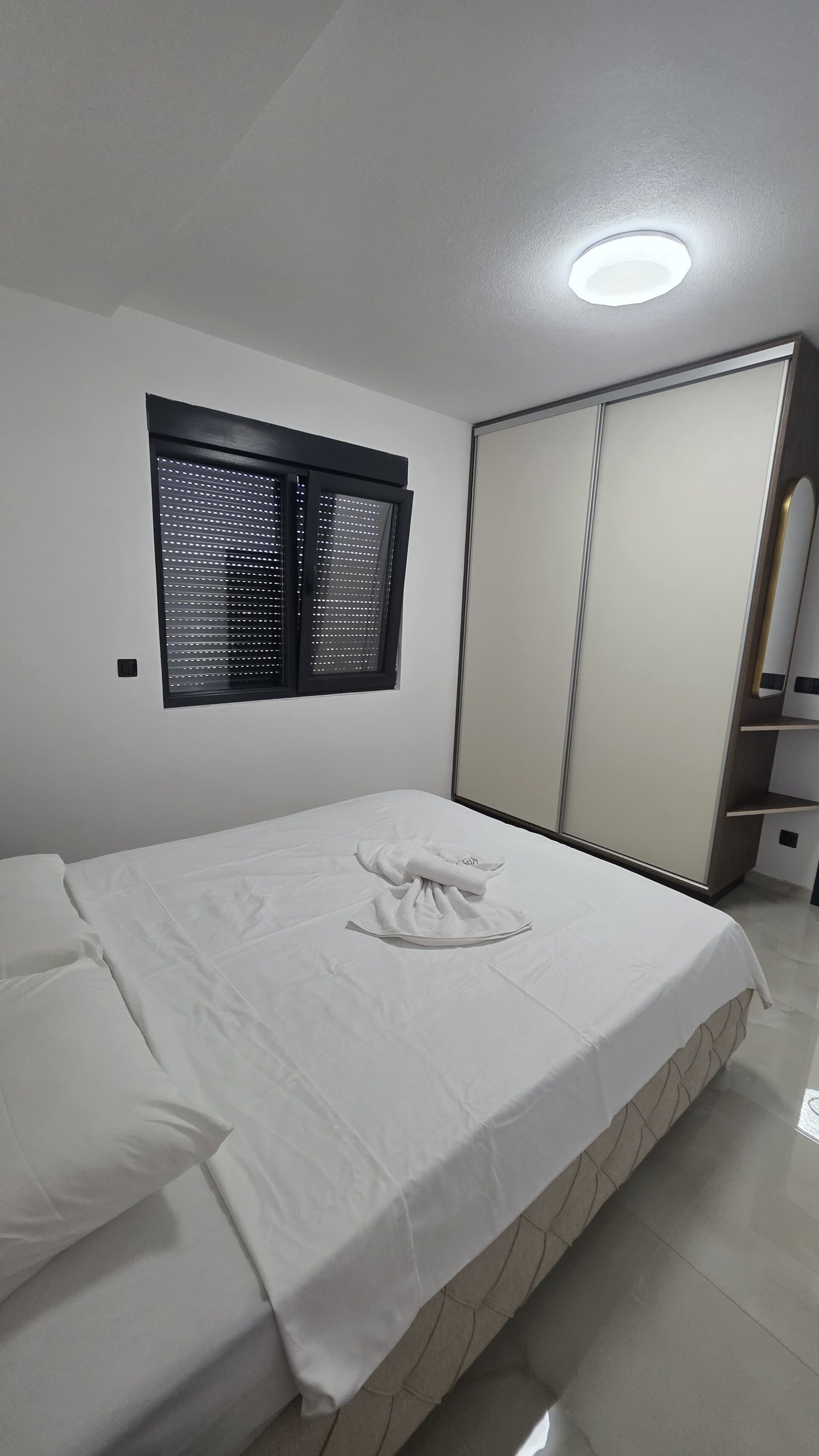 A bedroom with a bed, a window with blinds, a wardrobe, and a ceiling light.