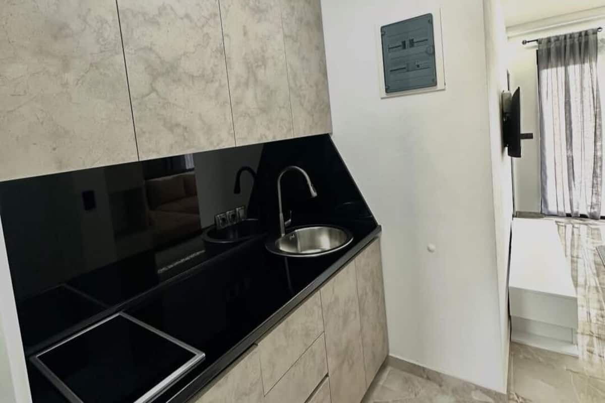 Une cuisine avec un plan de travail noir, un évier et des placards. Il y a une prise de courant sur le mur et une fenêtre avec des rideaux.