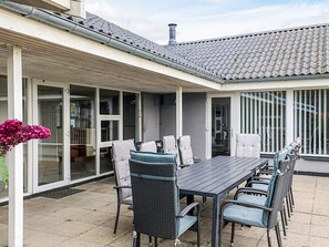 Une terrasse extérieure avec une table et des chaises, un espace salon abrité et une vue sur l’intérieur à travers des portes vitrées.