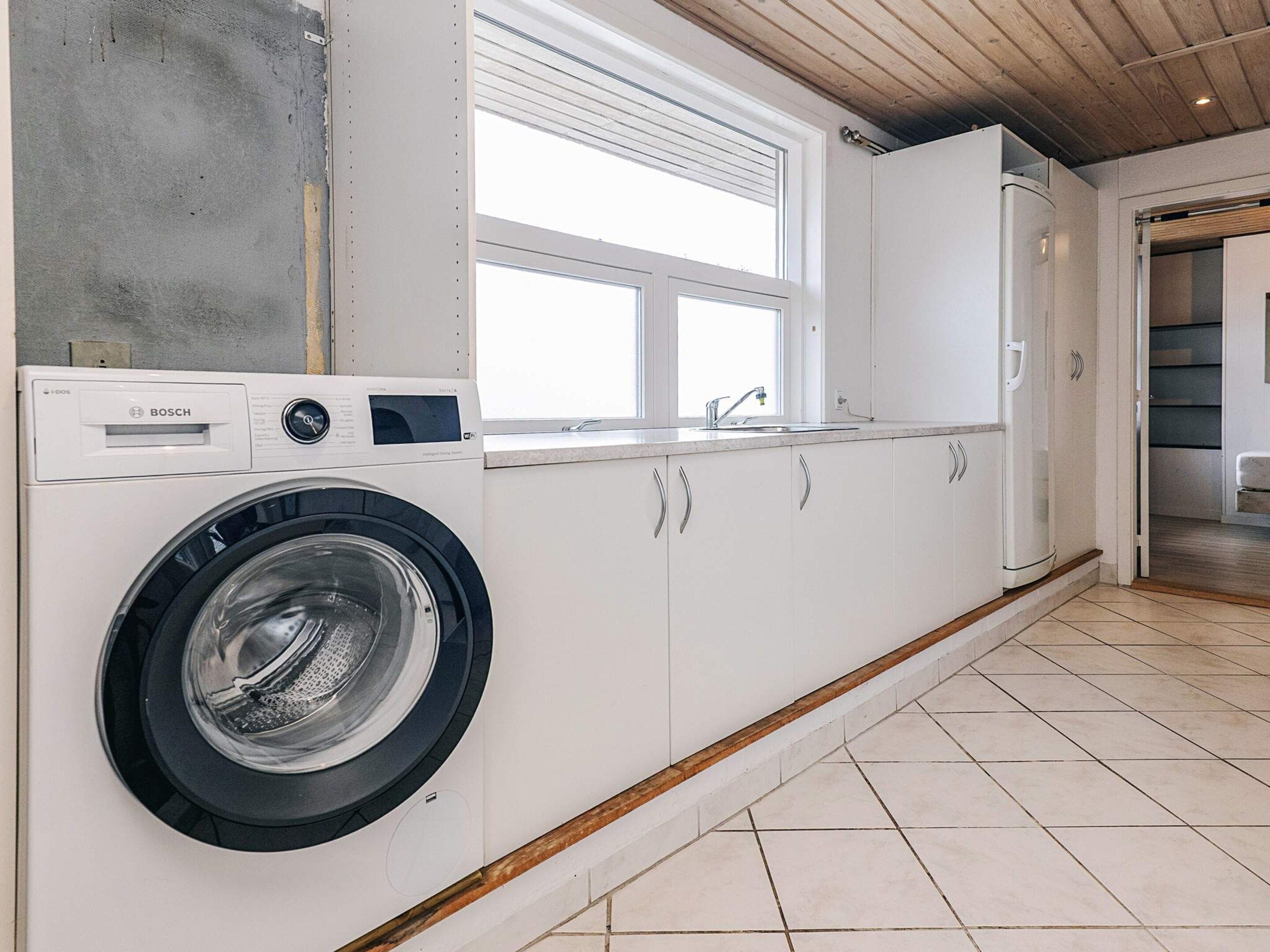 A compact laundry room with a Bosch washing machine, white cabinets, a window, and a wooden ceiling.
