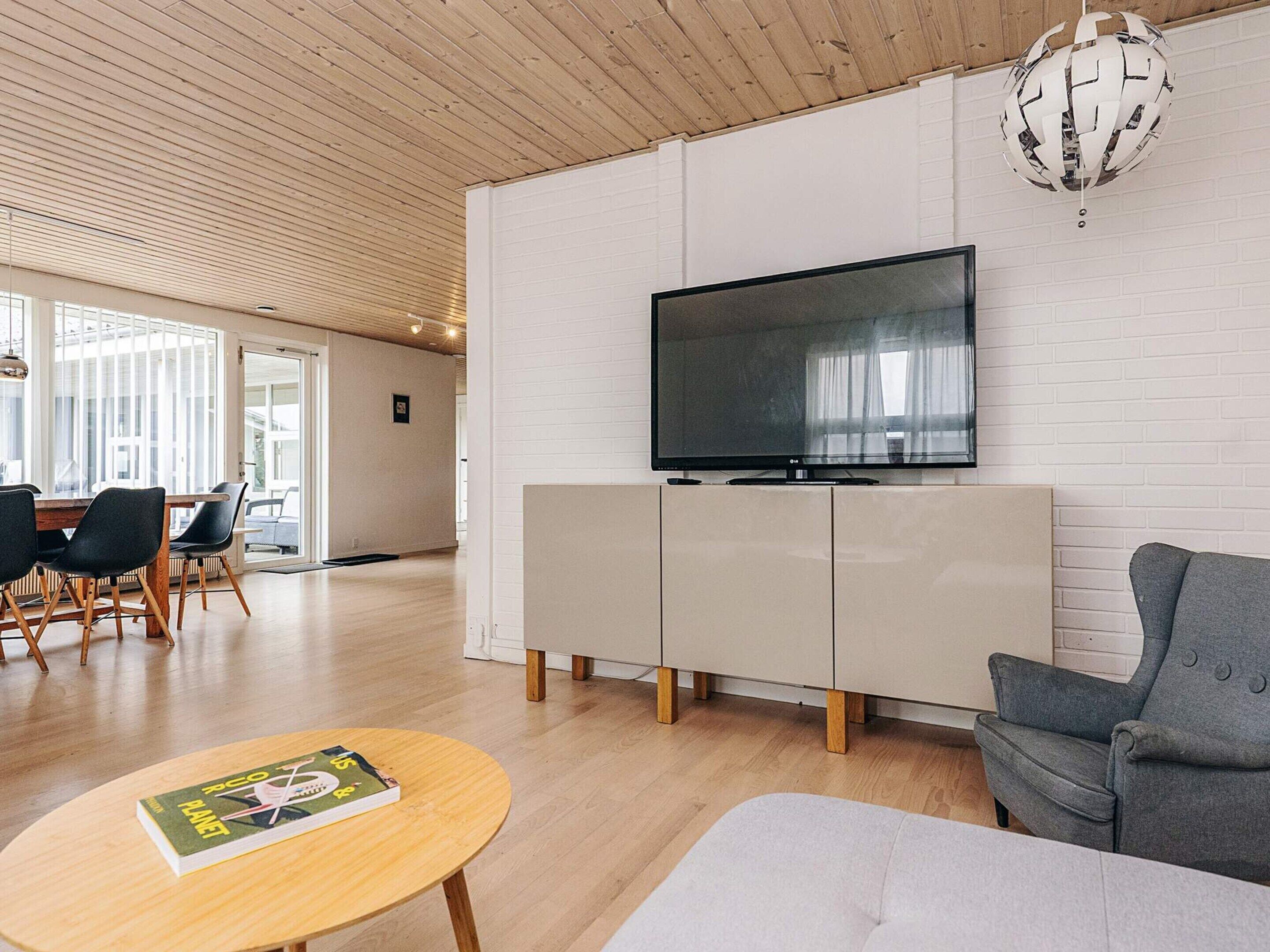 A modern living room with a flat-screen TV, a grey sofa, a wooden coffee table, and a dining area with chairs and a table.