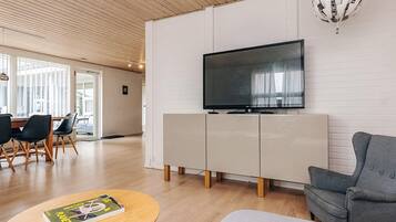 A modern living room with a flat-screen TV, a grey sofa, a wooden coffee table, and a dining area with chairs and a table.