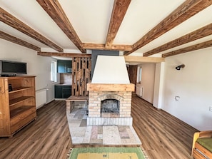 A living room with exposed wooden beams, a fireplace, a television, and a kitchen area.