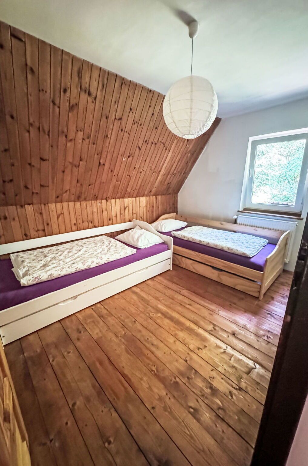 A room with two wooden beds, a ceiling fan, and a window with a view of trees.