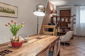Ein Essbereich mit Holztisch, Stühlen, einem roten Korb mit Brot, einer Blumenvase, einer Stehlampe, einem Bücherregal und einer Tür.