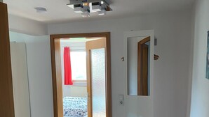 Huoneessa on puulattia, kuviollinen päiväpeite sängyllä, lasipaneelilla varustettu ovi ja pieni valkoinen kaappi, jonka päällä on vaaleanpunainen koristetyyny.