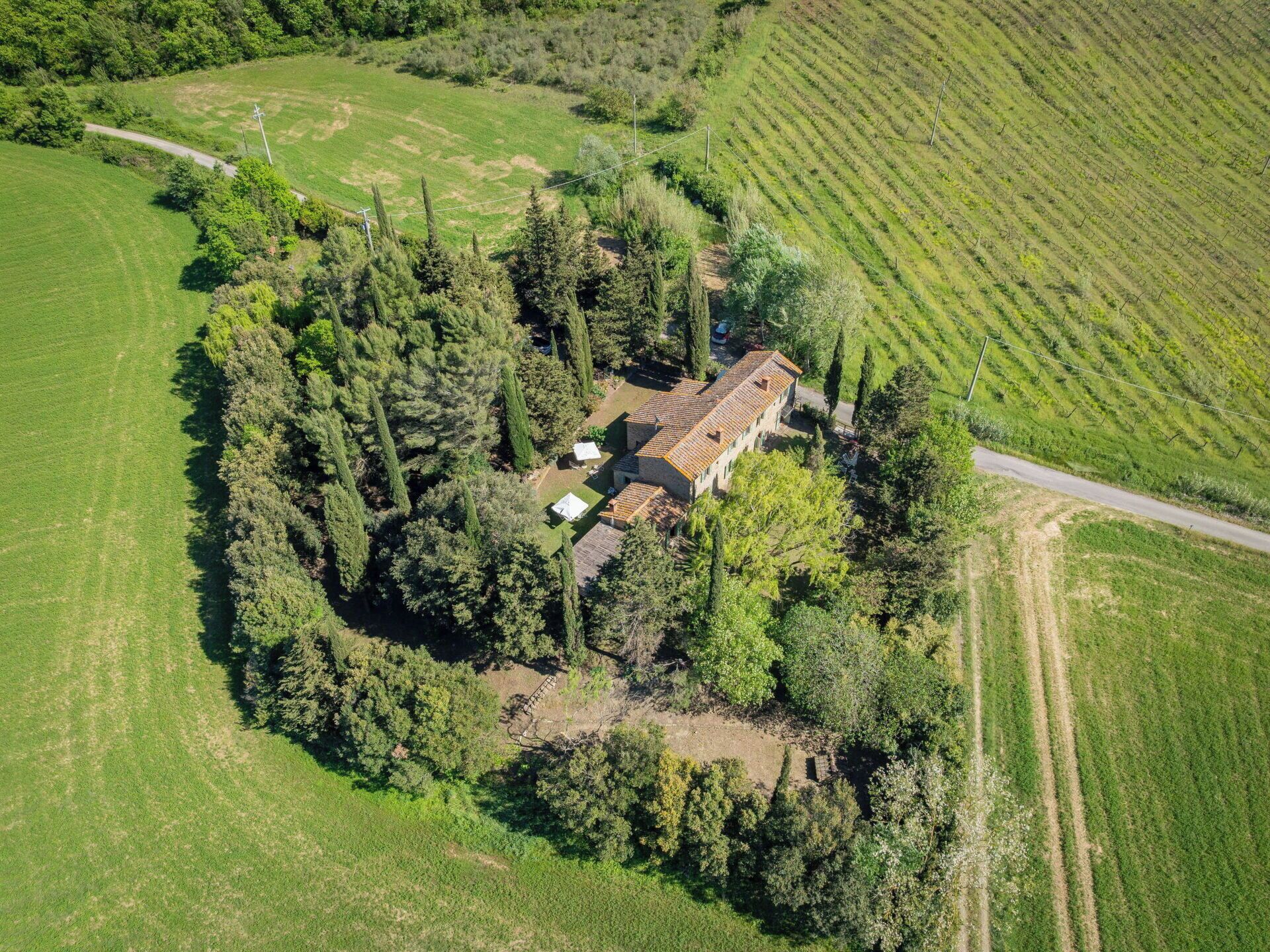 Vue aérienne d’une propriété rurale, avec une maison entourée d’une forêt dense, une route sinueuse et des champs agricoles adjacents.