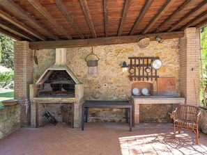 Un horno de ladrillo al aire libre con chimenea, un muro de piedra, una rejilla metálica con vajilla y un banco.