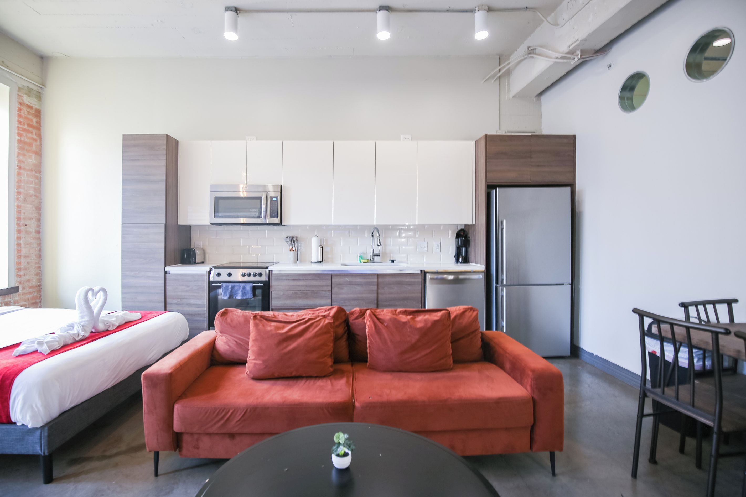 A compact hotel room with a bed, a red sofa, a small table, a kitchen area with a microwave, stove, and refrigerator, and a dining area with a chair.