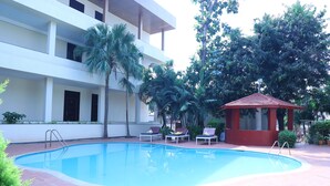 Una piscina con sillas de descanso, un pequeño pabellón y un edificio de varios pisos con balcones.