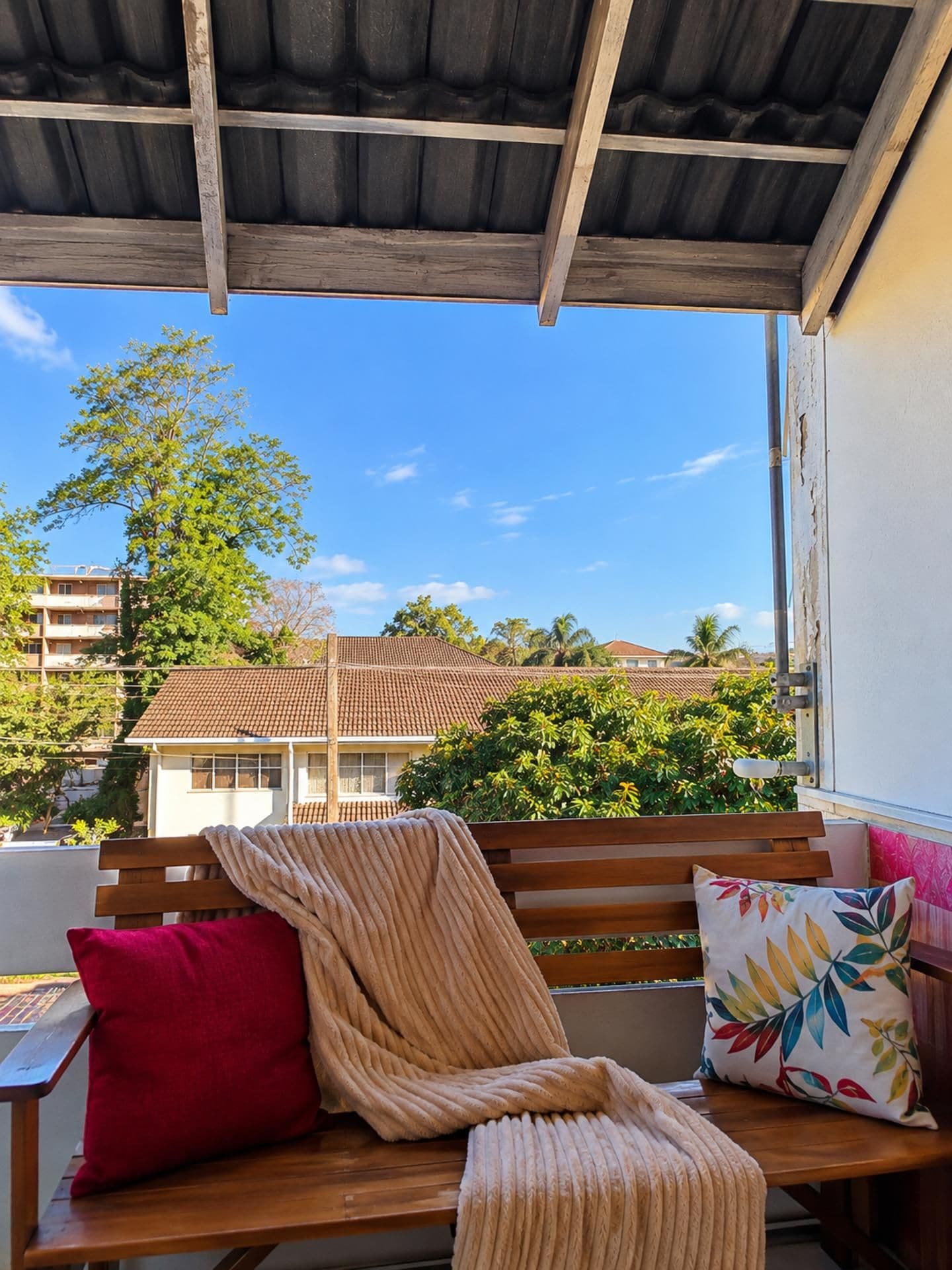 Un banco de madera con cojines y una manta, con vistas a una zona residencial con una casa de techo de tejas y vegetación.