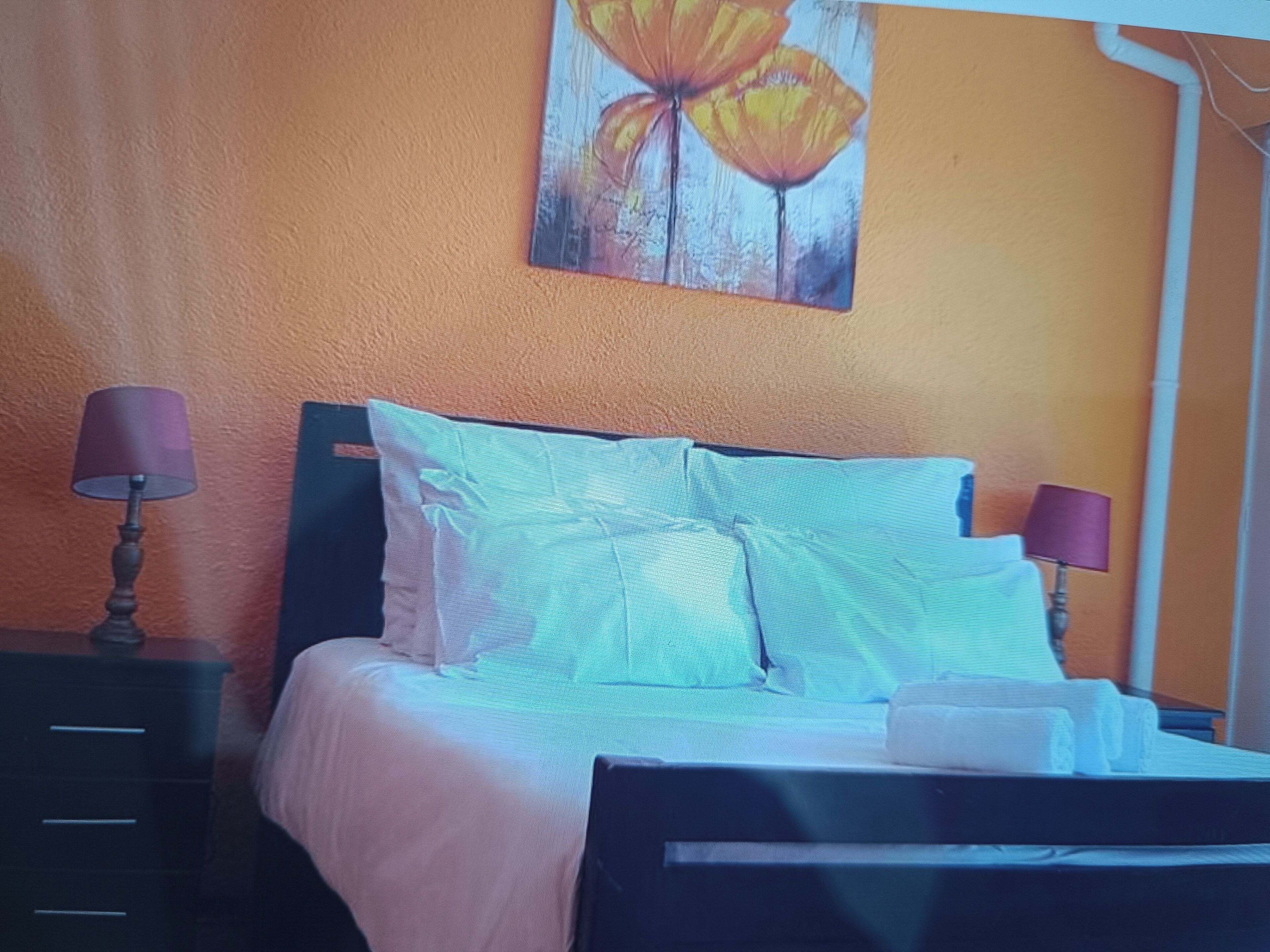 Una cama con sábanas blancas y dos almohadas, una mesita de noche con una lámpara, y un cuadro de flores en la pared.