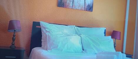 Una cama con sábanas blancas y dos almohadas, una mesita de noche con una lámpara, y un cuadro de flores en la pared.