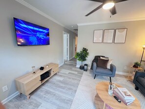 En moderne stue med et fladskærms-tv, en trækommode, en grå sofa, et sofabord og en potteplante.