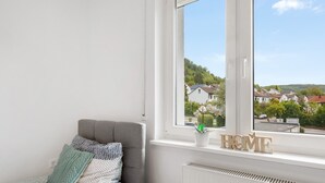 Une chambre à coucher avec un lit, des oreillers, une fenêtre, un radiateur et une pancarte « MAISON » posée sur le rebord de la fenêtre.
