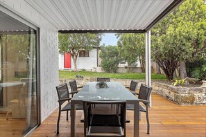 Une terrasse extérieure avec une table rectangulaire et des chaises, une terrasse en bois et une porte vitrée donnant sur un mur de briques.