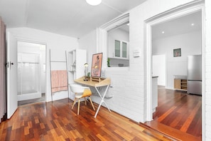 Un intérieur moderne avec un plancher en bois, des murs en briques blanches, un bureau avec une chaise et un coin cuisine visible à travers une porte ouverte.