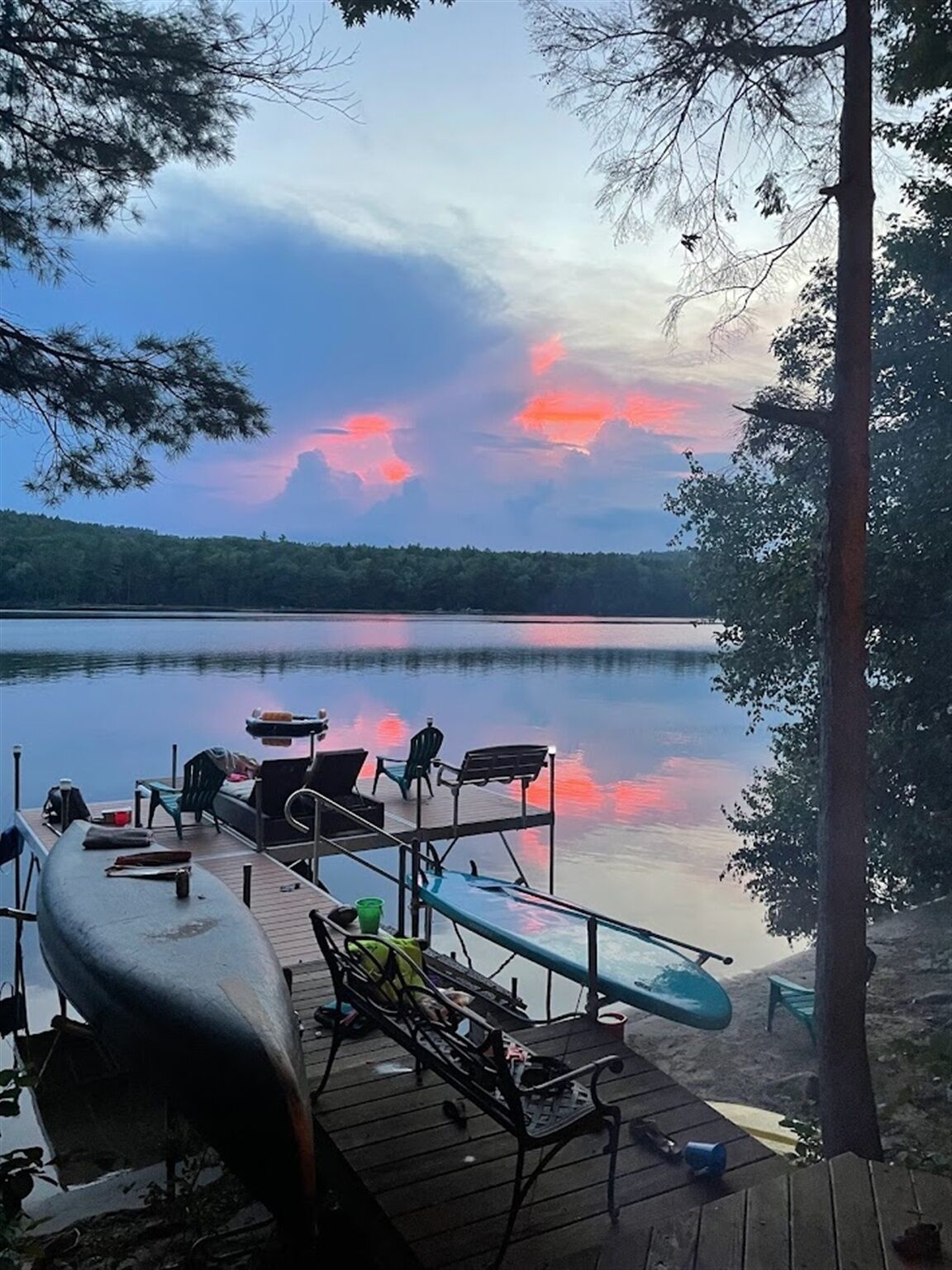 A dock with chairs and a kayak, overlooking a calm lake with trees and a sunset.