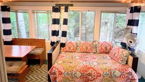 Interior de una cabaña con una cama, una mesa y un ventanal con vistas a un bosque.