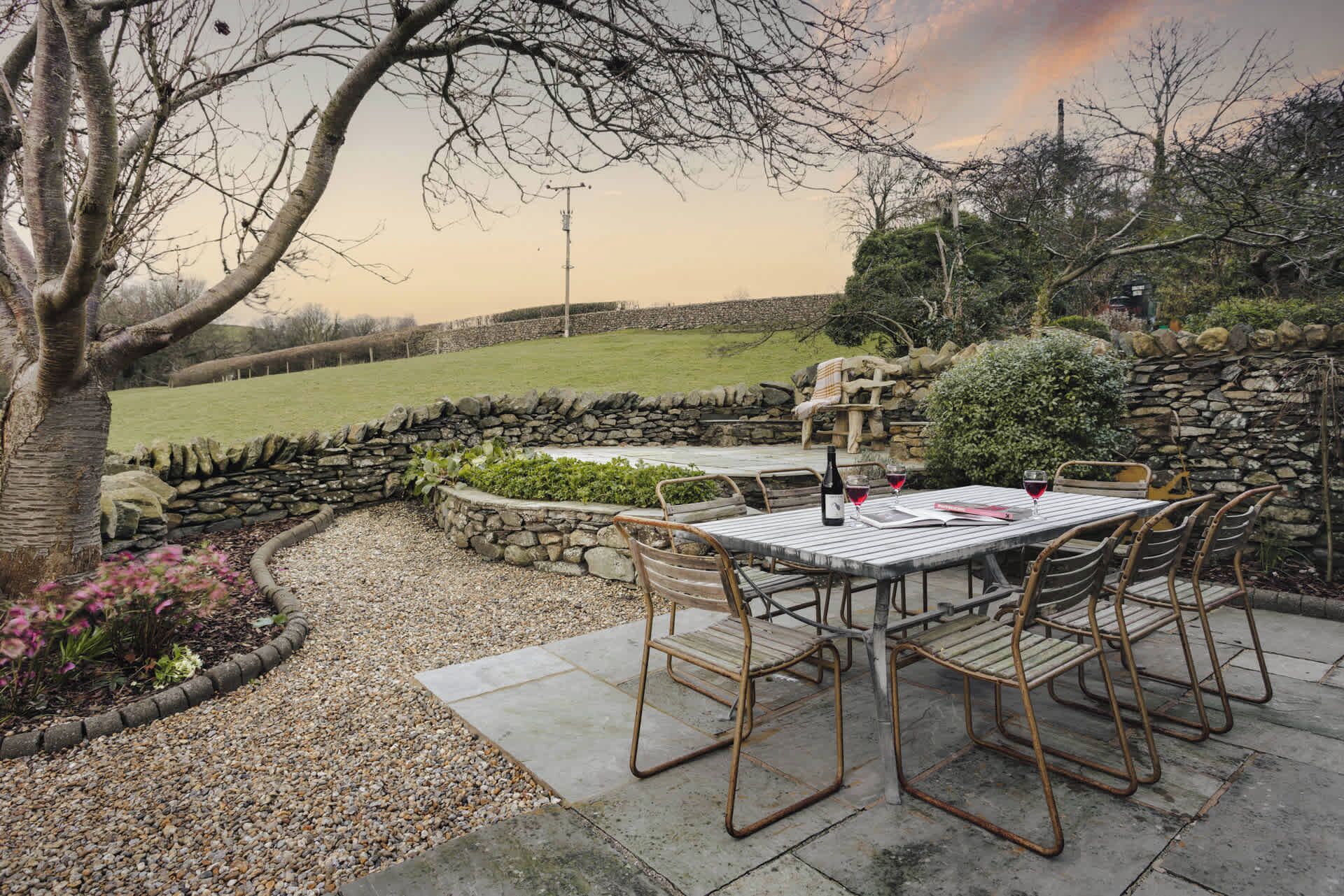 An outdoor dining area with a stone wall, a table with chairs, and a view of the countryside.