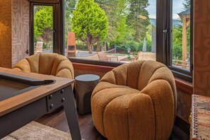 A room with two upholstered chairs, a small round table, and a view of a garden through large windows.
