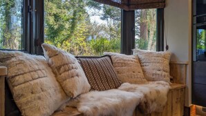 A wooden bench with fur cushions, large windows, and a view of trees. - Wyke View | Ridgestone (Bowness-on-Windermere)