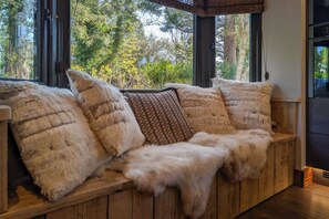 A wooden bench with fur cushions, large windows, and a view of trees.