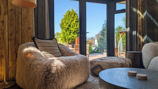 A cozy room with a large window, a fluffy chair, a round table, and a view of greenery outside. - Wyke View | Ridgestone (Bowness-on-Windermere)