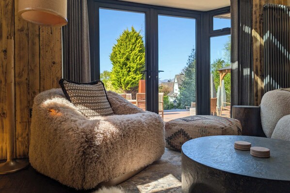 A cozy room with a large window, a fluffy chair, a round table, and a view of greenery outside.