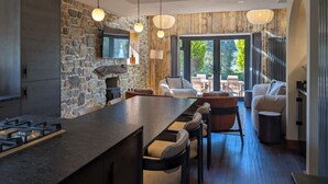 A modern kitchen with a stone wall, a large island, bar stools, and a dining area with a view. - Wyke View | Ridgestone (Bowness-on-Windermere)