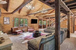 A log cabin interior with a wooden ceiling, a staircase, a fireplace, and a large window.
