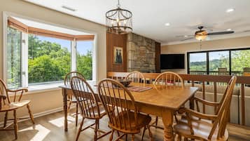A dining area with a large wooden table, six chairs, a chandelier, a stone wall, and a television mounted on the wall.