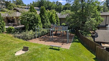 A playground with climbing structures and a bench, surrounded by a wooden fence and greenery.