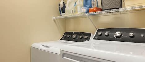 A front-loading washing machine with a control panel, a shelf above it holding laundry supplies, and a laundry basket with a towel.