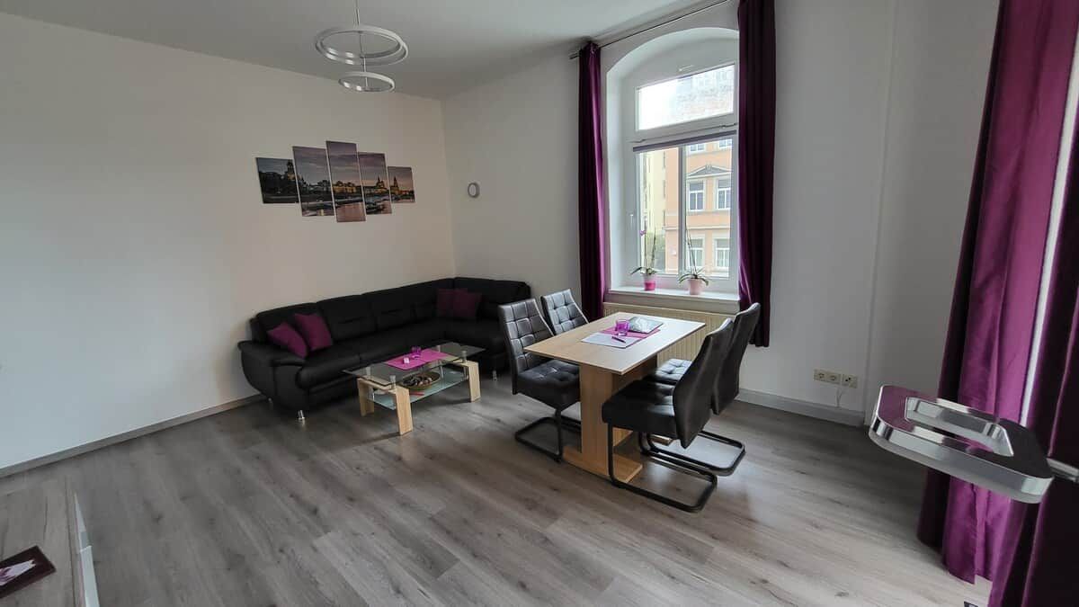 A modern living room with a black sofa, a dining table with chairs, a small coffee table, and a window with curtains.