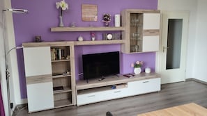 Ein Wohnzimmer mit Fernseher, Holzregalen, einer Vitrine und einer Tür.