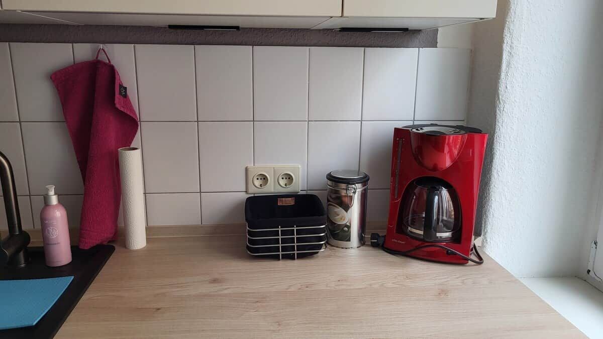Un comptoir de cuisine avec une machine à café, un bocal, un panier noir, une bouteille rose et une serviette rouge.