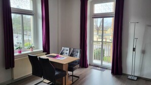 Habitación con una mesa, sillas, un radiador, una ventana con cortinas y vistas a edificios.