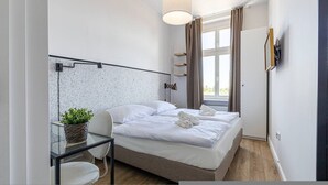 A modern bedroom with a bed, a small table with a plant, a chair, a bedside lamp, and a window with curtains. - Verde 5 Gda sk Pok j z Azienk Blisko Centrum (Gdańsk)