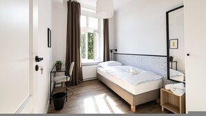 Une chambre à coucher comprenant un lit, une chaise, une petite table, un radiateur, une fenêtre avec des rideaux et un miroir.
