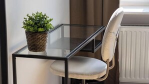Un bureau à plateau en verre, un pot de fleurs en osier, une chaise blanche rembourrée et un radiateur dans une pièce au plancher en bois et dotée d’une fenêtre avec des rideaux.