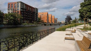 Un paseo fluvial con un puente, bancos y edificios modernos al fondo.