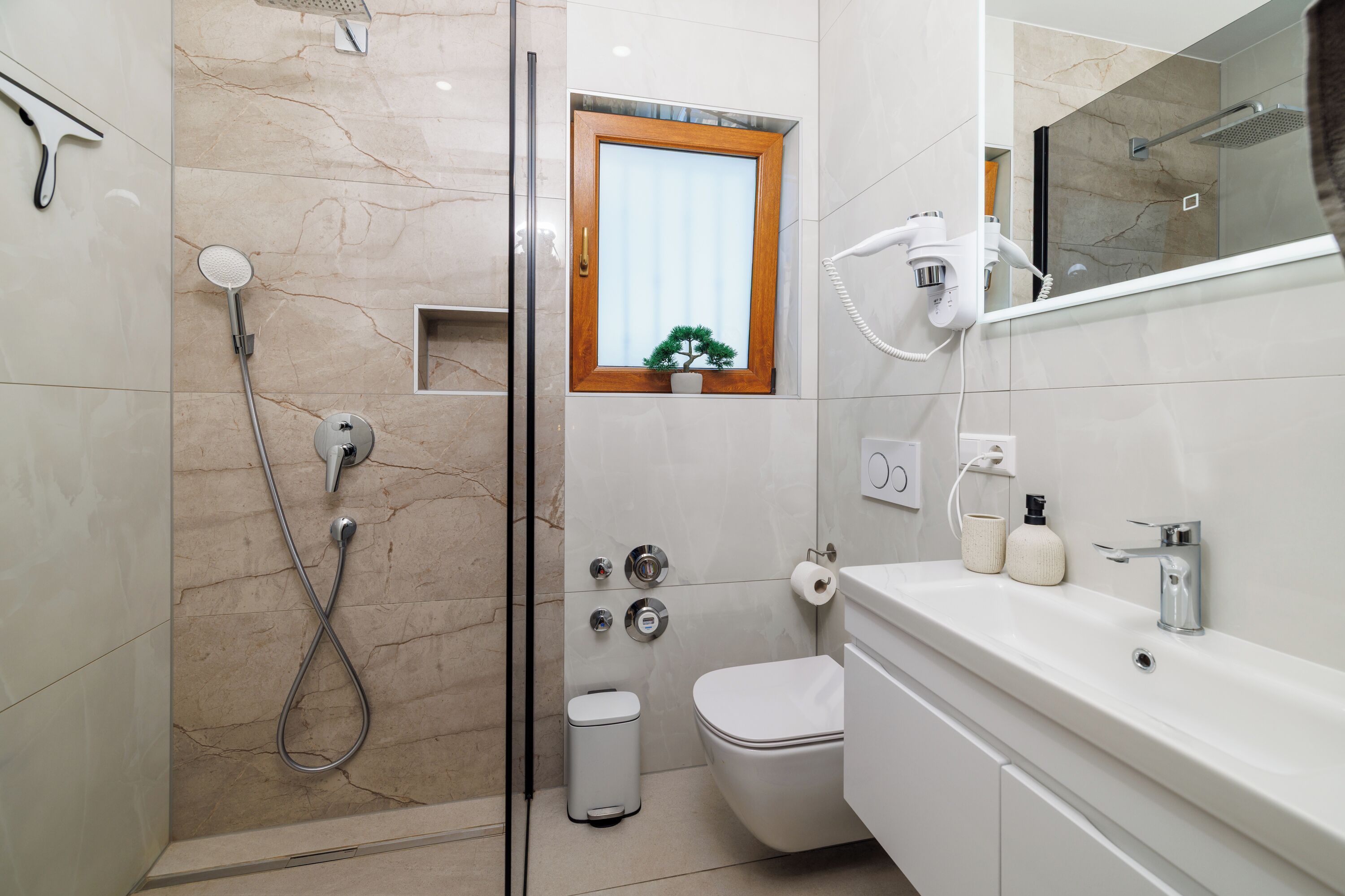 Un baño moderno con ducha, inodoro y lavamanos, con paredes de mármol y un pequeño ventanal con una planta.