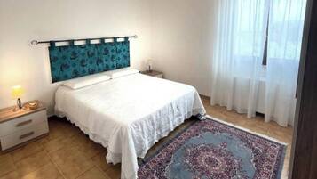 Un dormitorio con cama, mesita de noche, lámpara, cabecera estampada, ventana grande con cortinas y alfombra estampada.