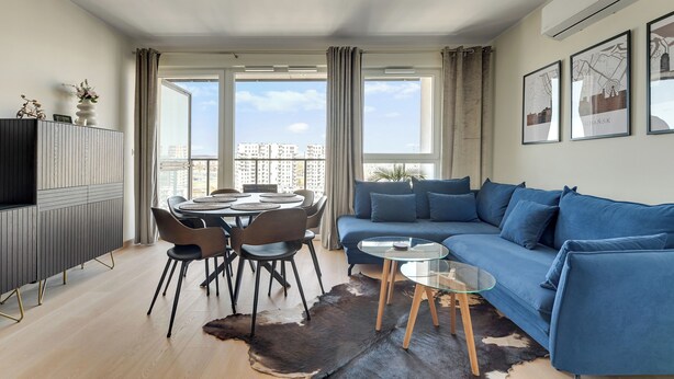 A modern living room with a blue sofa, a glass coffee table, a dining table with chairs, and a large window with curtains. - RentPlanet - Apartament Starowiejska (Gdańsk)