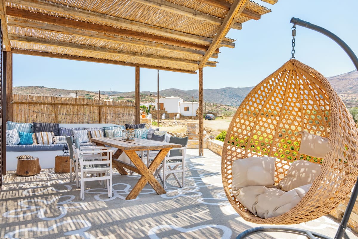 An outdoor seating area with a wooden table and chairs, a hanging chair, and a thatched roof structure.